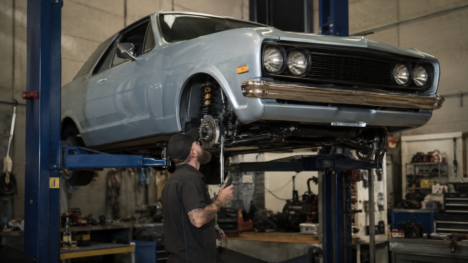 Car on a hoist in a workshop bay with front wheels off the ground and a technician inspecting the suspension
